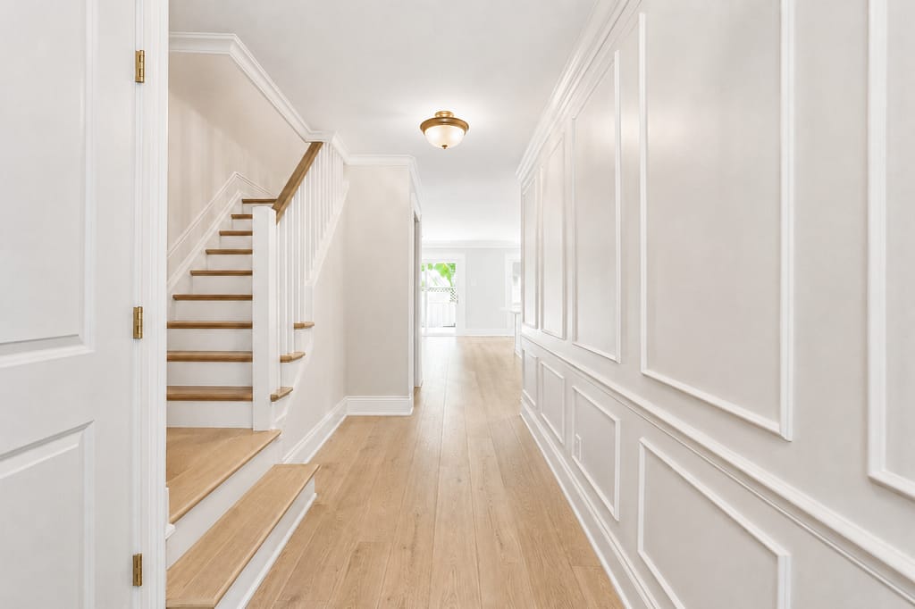 Finished hallway with wall paneling, baseboards, shoe moulding, crown moulding and a door with brushed brass hinges beside the staircase