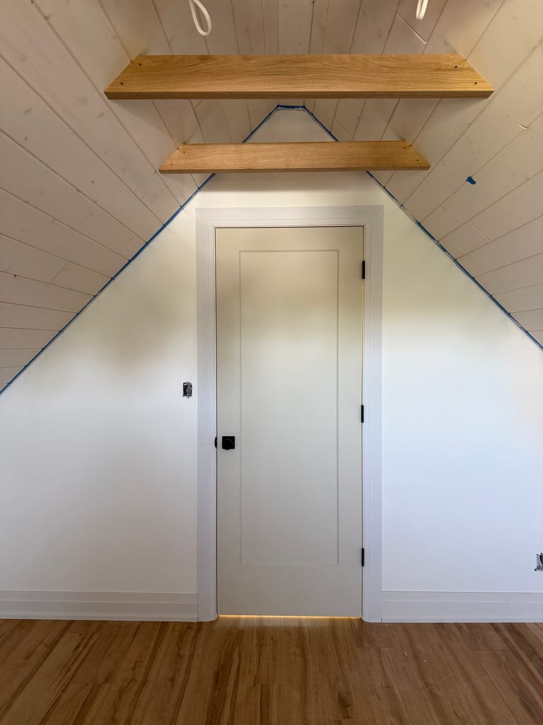 Interior attic door with casing, baseboards and wood shelves under a steep sloped ceiling