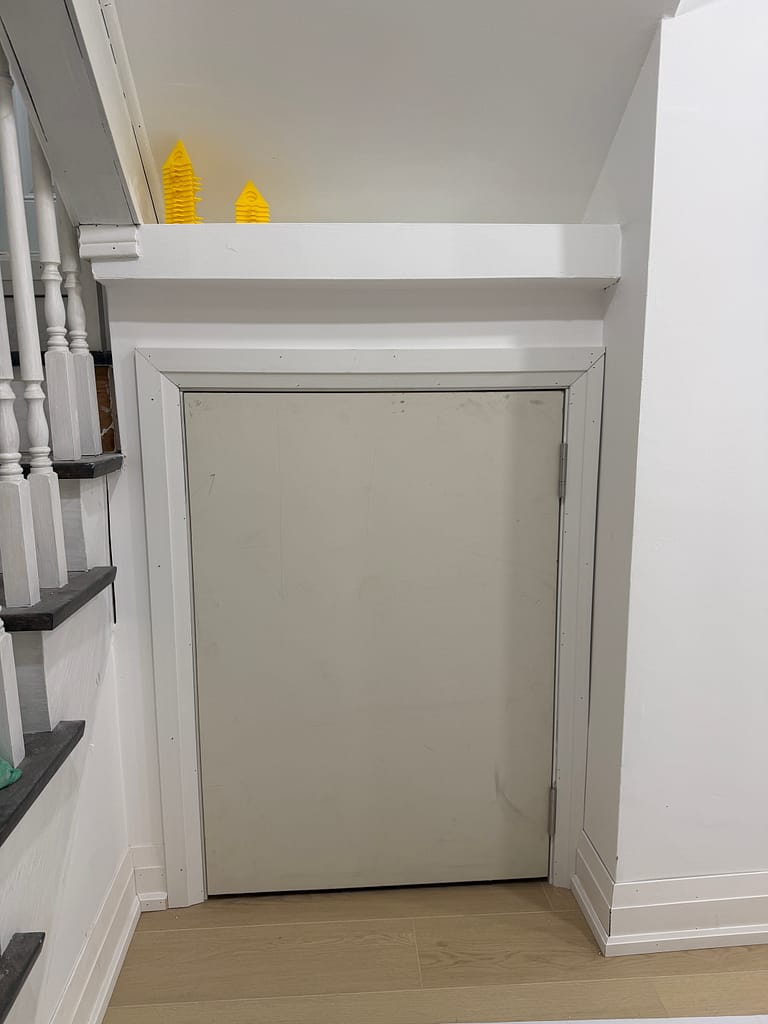 Custom basement storage door installed under stairs with casing and baseboard trim around a low opening.