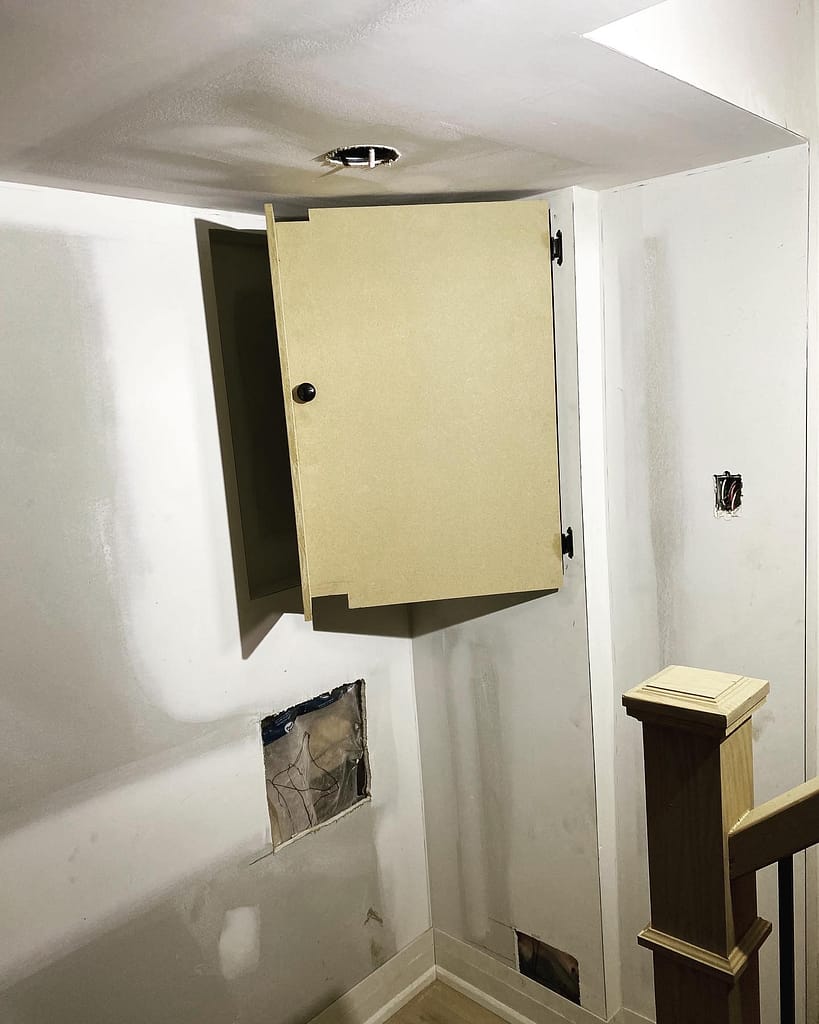An open custom-built wood wall panel enclosure hiding an old electrical fuse box seamlessly inside updated hallway trim.