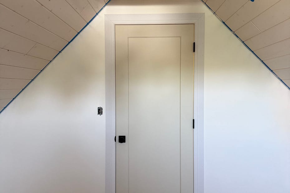 Interior attic door with casing, baseboards and wood shelves under a steep sloped ceiling
